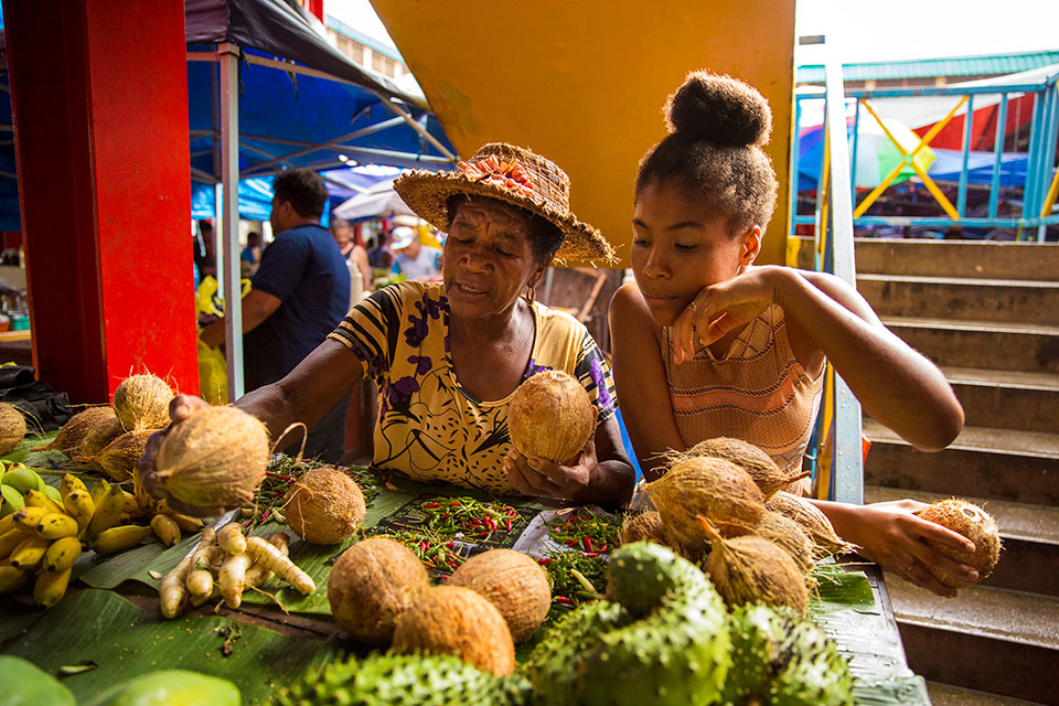 Mana Celestine (left) sells bananas, coconuts, papaya, chillis and soursop in the market in Victoria with the help of her granddaughter Anel. Photo: UN Women/Ryan Brown