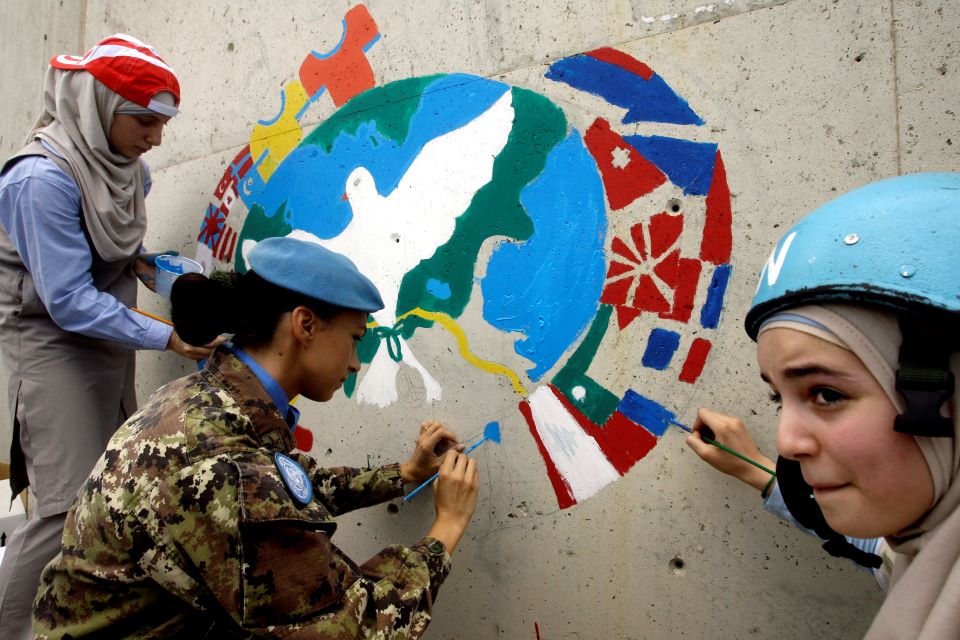 Three women paint a mural symbolizing peace on a wall. 